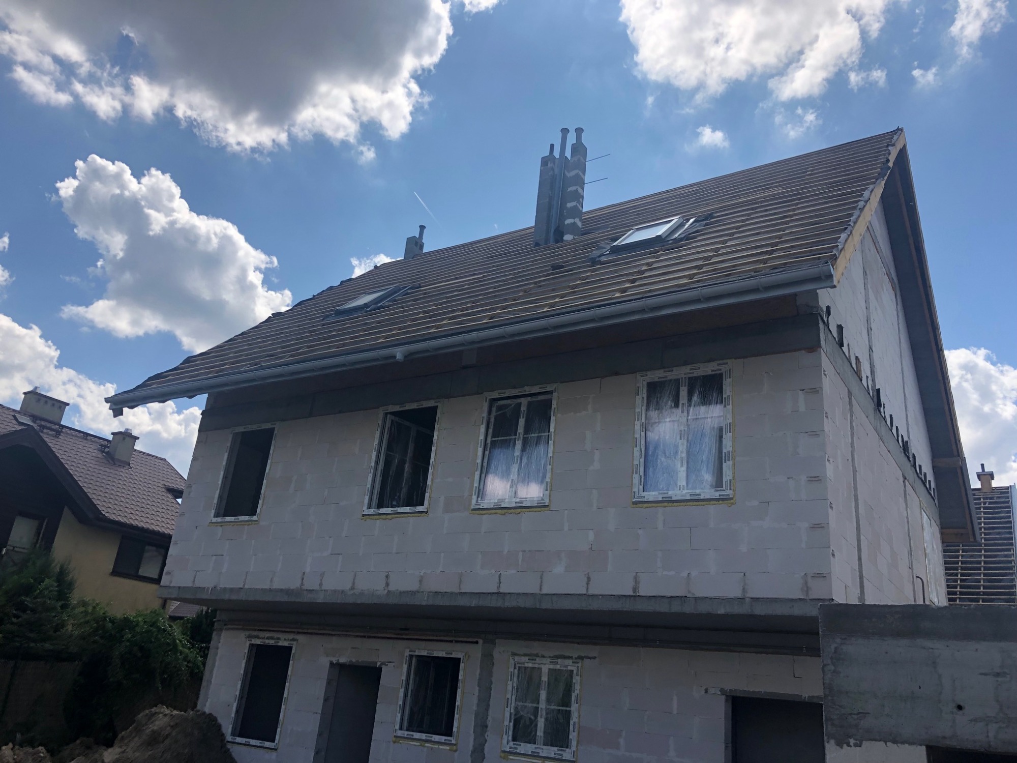 Budowa dwupiętrowego domu z białych bloczków, z ukończoną konstrukcją dachu pokrytą brązową dachówką, widoczne okna dachowe i kominy, w tle błękitne niebo z chmurami.