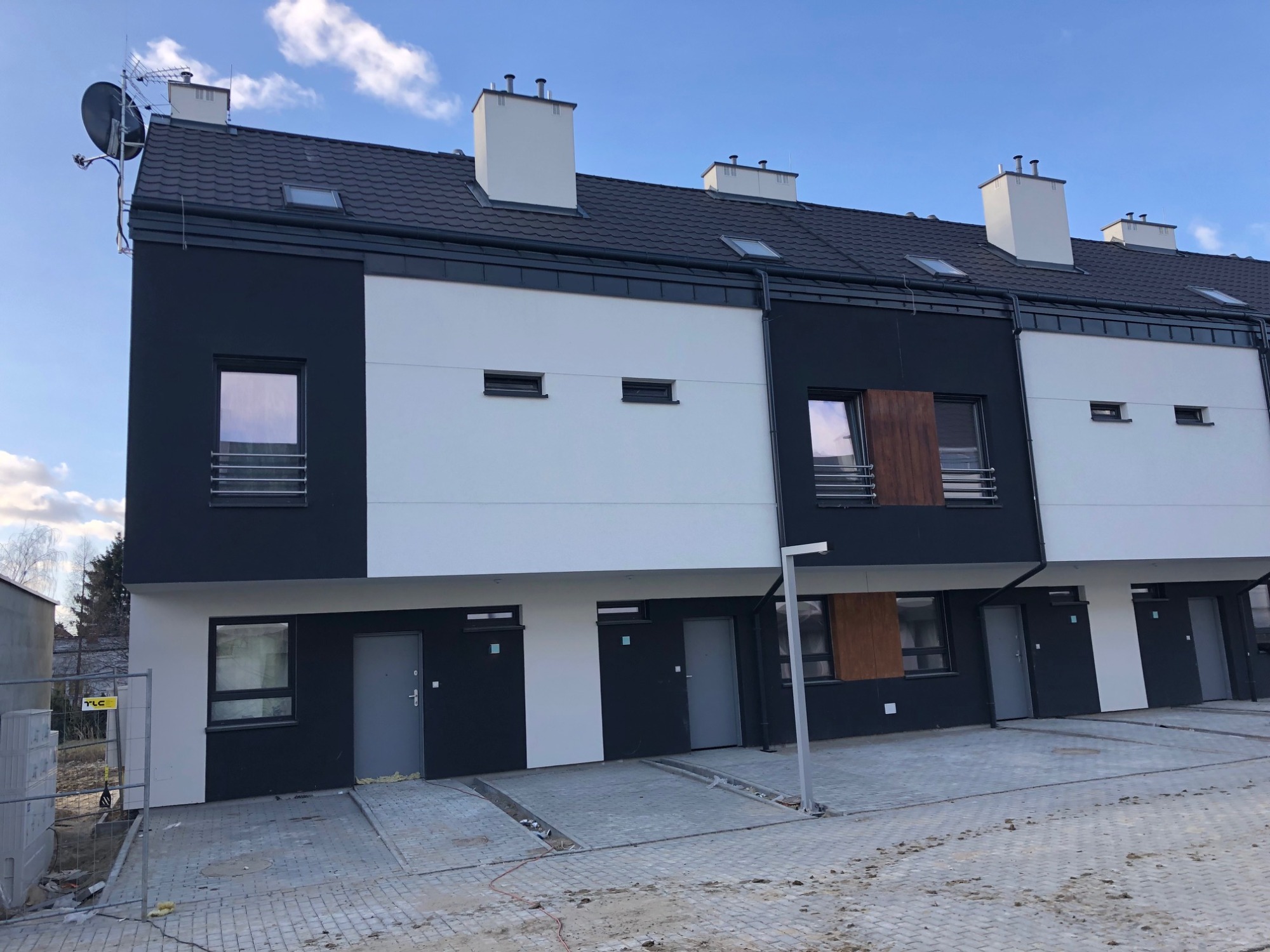 Nowoczesne segmenty mieszkalne w zabudowie szeregowej, z czarno-białą elewacją, grafitowym dachem i widoczną anteną satelitarną na jednym z budynków, teren wokół w trakcie prac wykończeniowych.