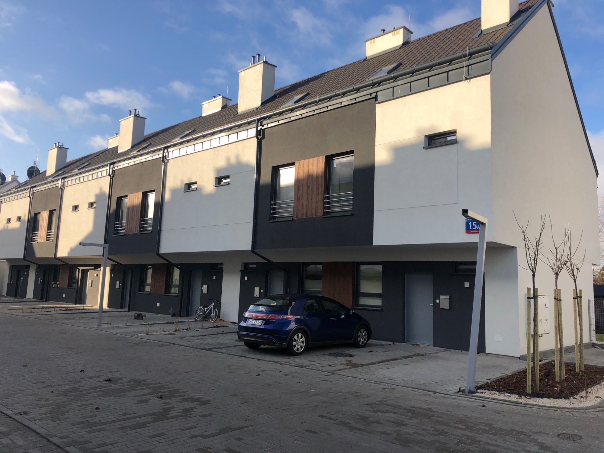 Nowoczesne szeregowe domy z ciemnoszarą dachówką, białymi ścianami i ciemnymi elementami elewacji, zaparkowany niebieski samochód i rower, ulica z kostki brukowej, błękitne niebo z chmurami w tle.