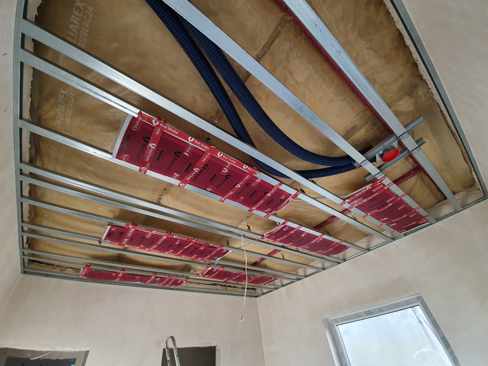 Montaż folii grzewczej Red Snake na suficie podwieszanym we Wrocławiu. Widoczne profile konstrukcyjne, izolacja, niebieskie rury instalacyjne i fragment drabiny.