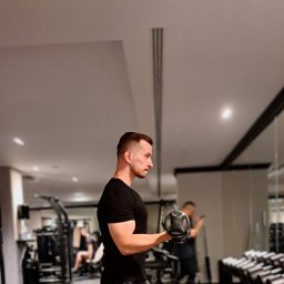 BanyGym Illegal Studio - Mężczyzna w czarnej koszulce ćwiczy biceps z hantlą 12 kg w siłowni, w tle widoczne maszyny do ćwiczeń i lustro.