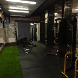 BanyGym Illegal Studio - Wnętrze siłowni z czarną gumową podłogą, sztuczną trawą, sprzętem do ćwiczeń, widocznymi cegłami i zegarem cyfrowym na ścianie.