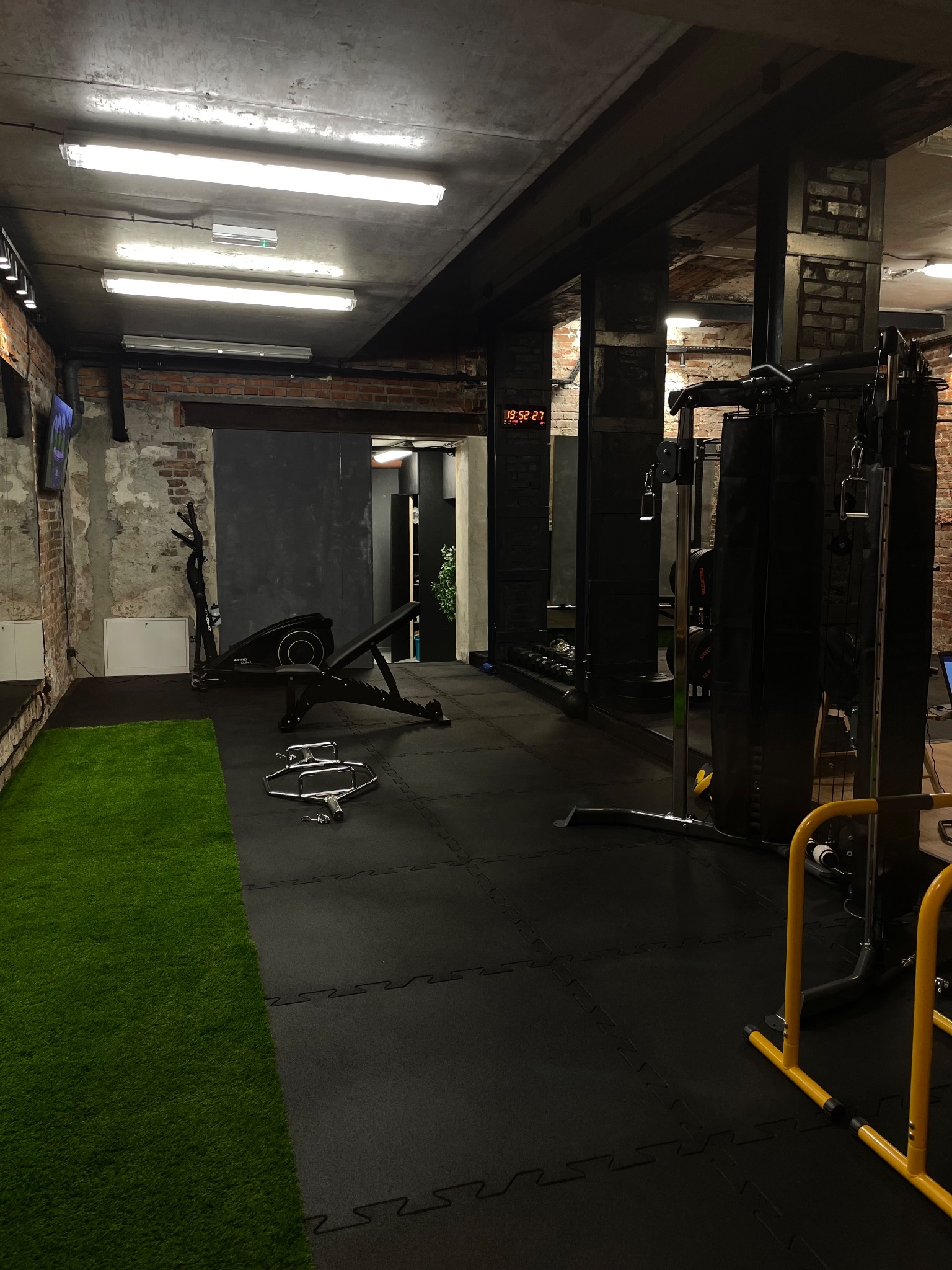Wnętrze siłowni z czarną gumową podłogą, sztuczną trawą, sprzętem do ćwiczeń, widocznymi cegłami i zegarem cyfrowym na ścianie.