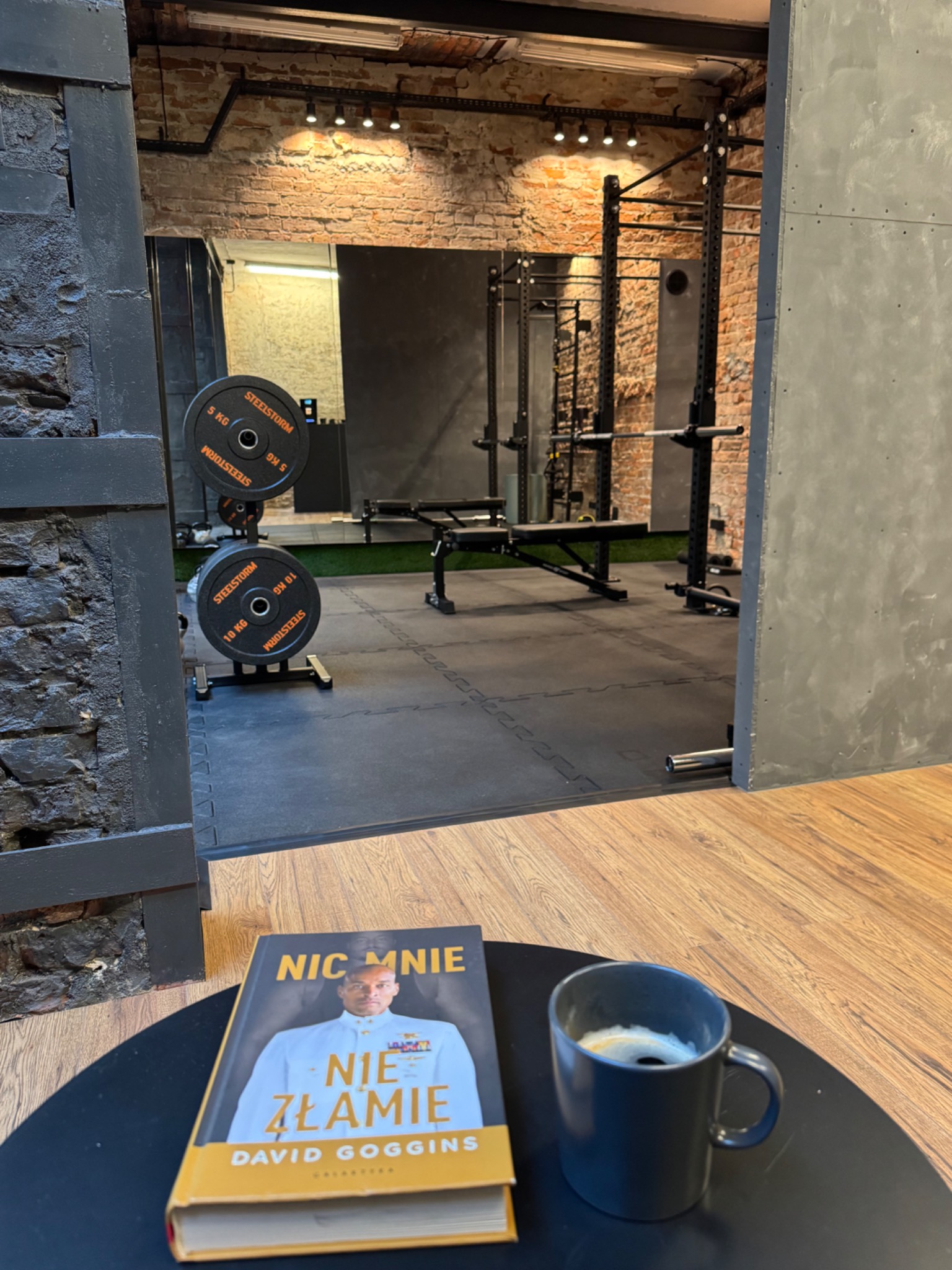 Książka Davida Gogginsa 'Nic mnie nie złamie' i filiżanka kawy na stoliku w tle siłowni z ceglaną ścianą i sprzętem do ćwiczeń.