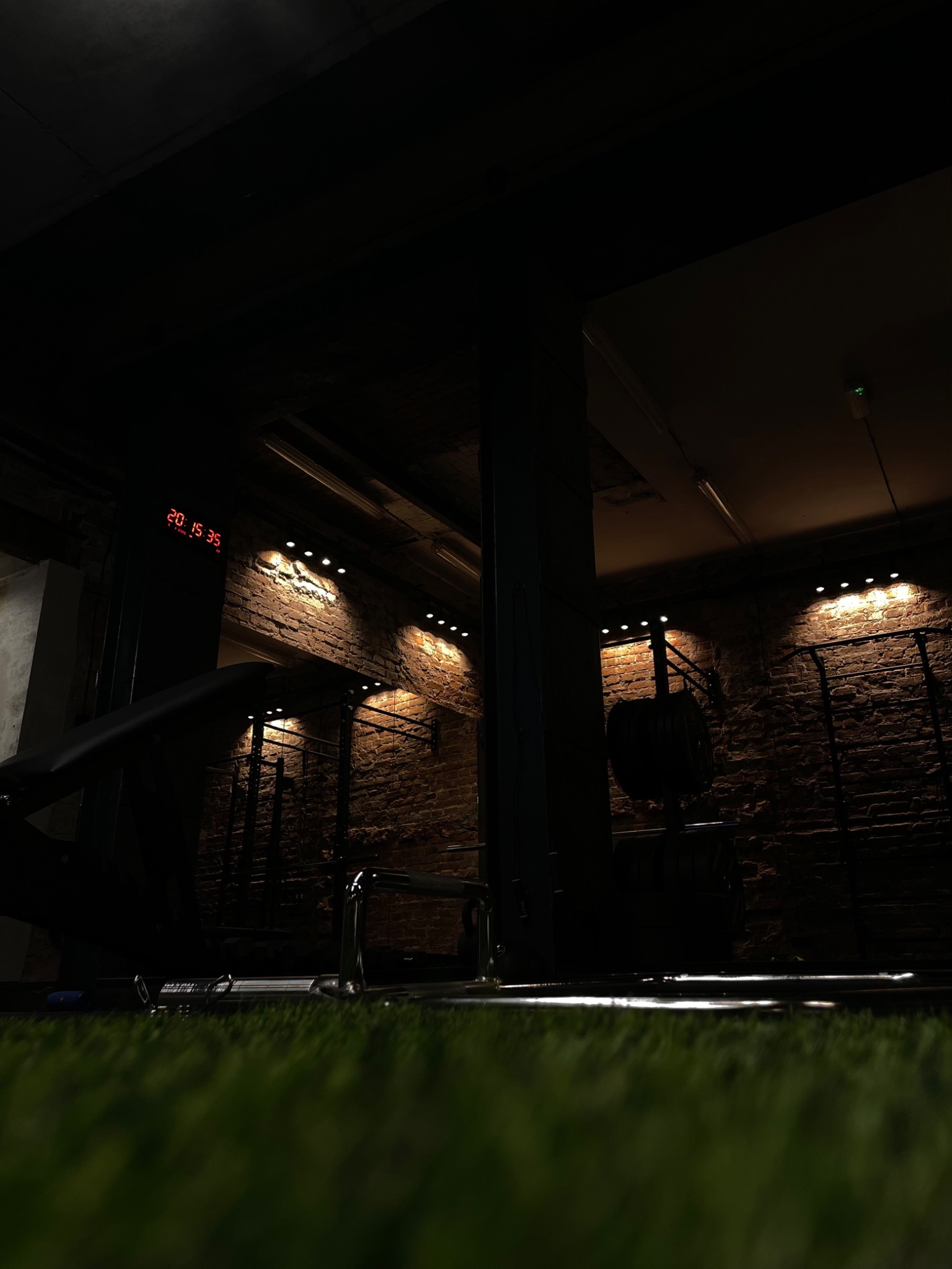 Ciemne wnętrze siłowni z ceglanymi ścianami oświetlonymi punktowymi lampami, widoczne urządzenia do ćwiczeń i sztuczna trawa na podłodze w pierwszym planie, zegar elektroniczny wyświetla godzinę 20...