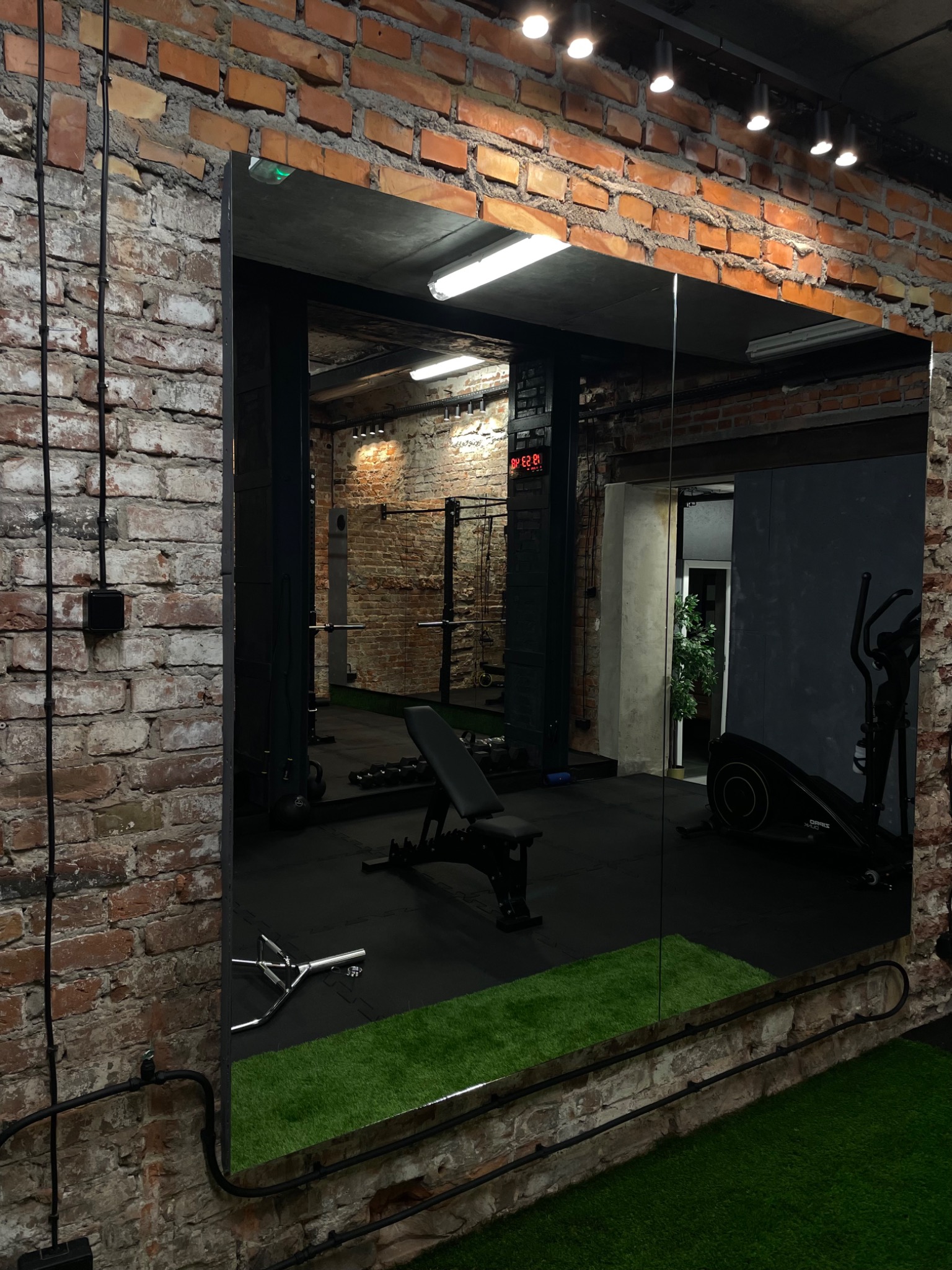 Widok na siłownię odbity w lustrze, z ceglanymi ścianami, czarnym sprzętem do ćwiczeń, sztuczną trawą i widocznym zegarem cyfrowym.