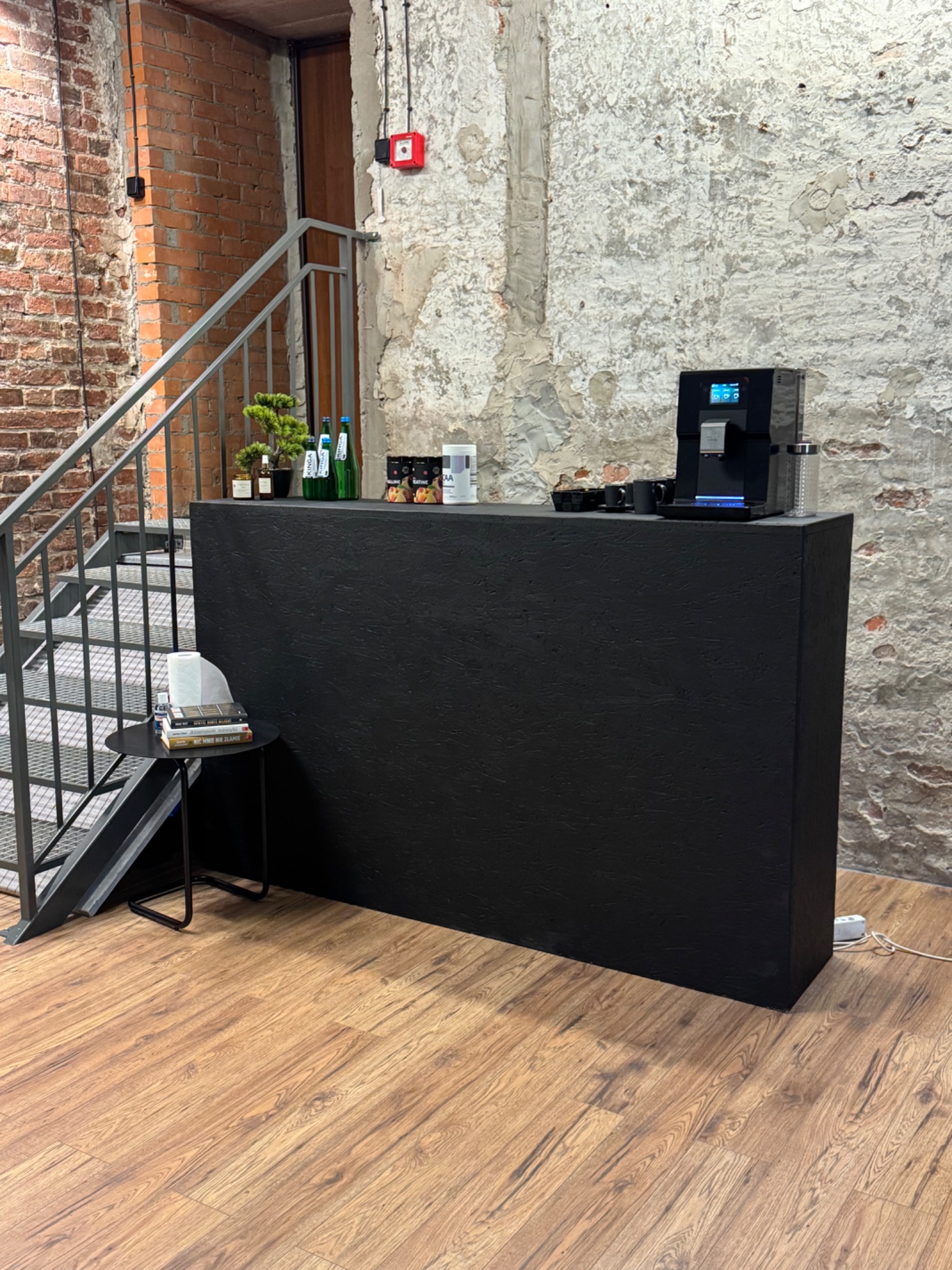 Czarny bar z ekspresem do kawy, suplementami diety i bonsai na tle surowej, ceglanej i otynkowanej ściany, obok schodów metalowych i stolika z książkami.