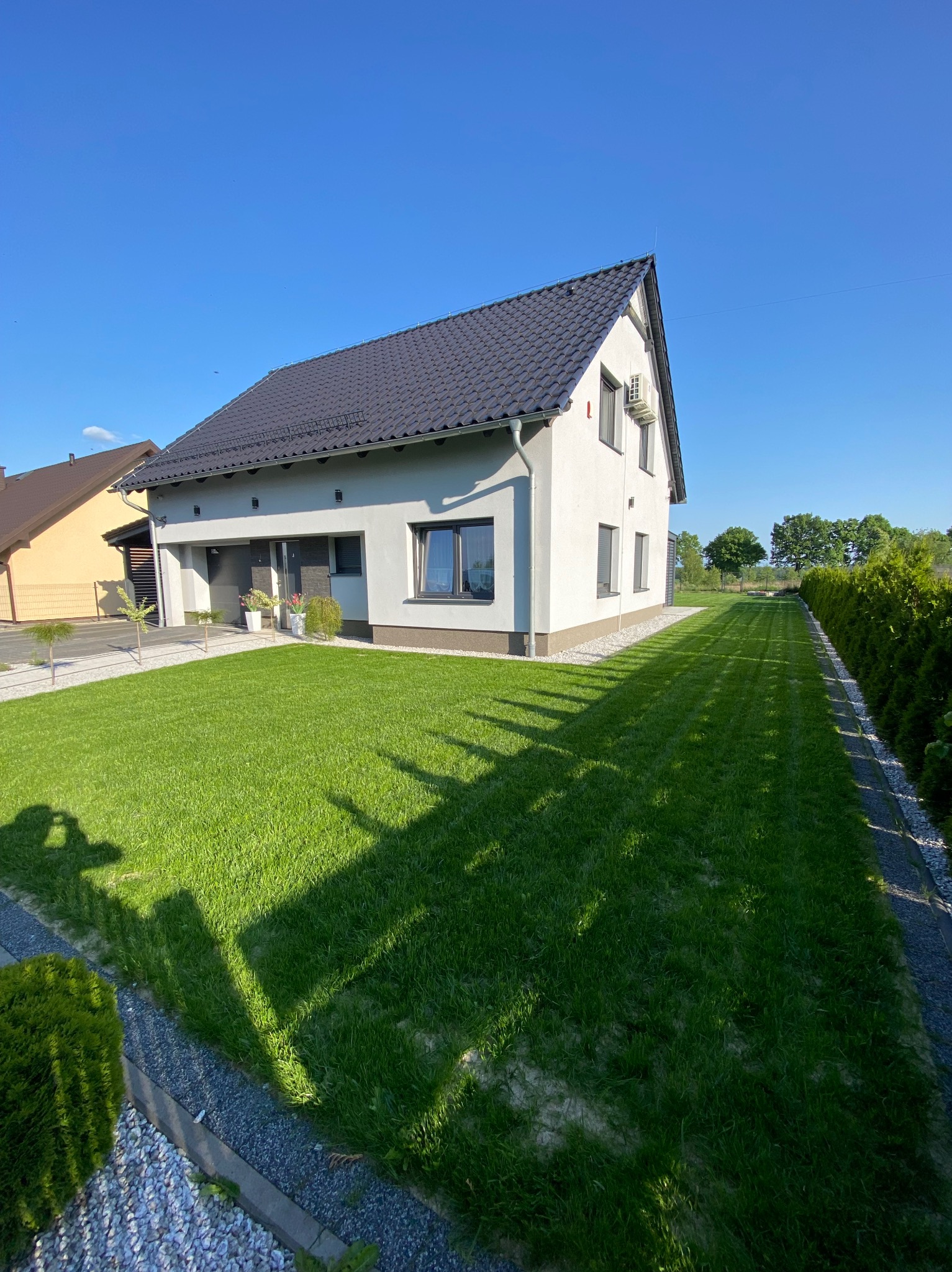 Nowoczesny dom jednorodzinny z ciemną dachówką i idealnie przystrzyżonym trawnikiem, wykończony ozdobnym kamieniem i żywopłotem, w słoneczny dzień.