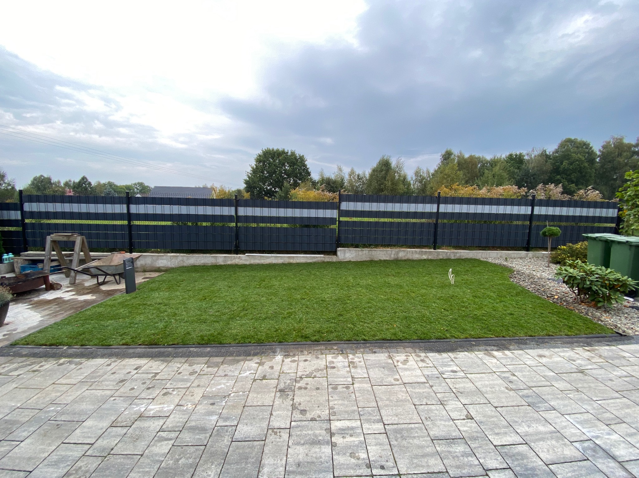 Świeżo ułożony trawnik z rolki przylegający do szarej kostki brukowej, w tle czarne ogrodzenie panelowe i pochmurne niebo.