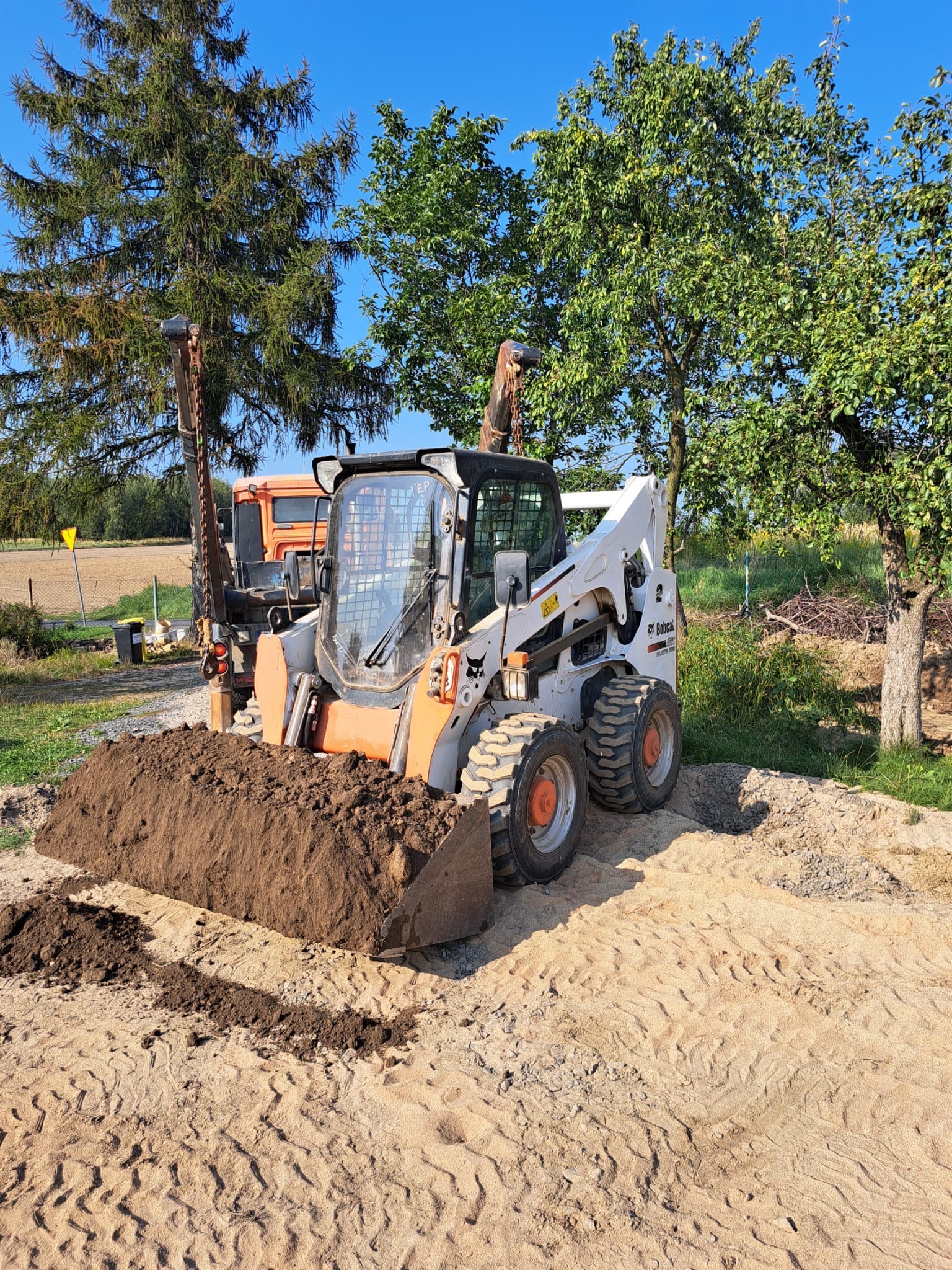Bobcat z pełną łyżką ziemi na piaszczystym terenie, w tle drzewa i częściowo widoczny pomarańczowy samochód ciężarowy.