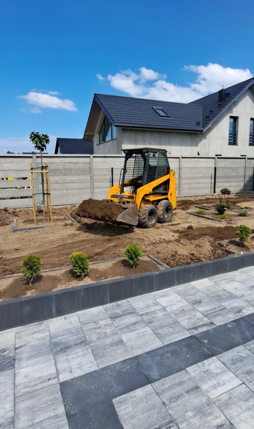 Żółta miniładowarka przewozi ziemię na terenie nowo zakładanego ogrodu z niskimi krzewami i świeżo posadzonym drzewem, w tle nowoczesny dom z szarą elewacją i grafitowym dachem.
