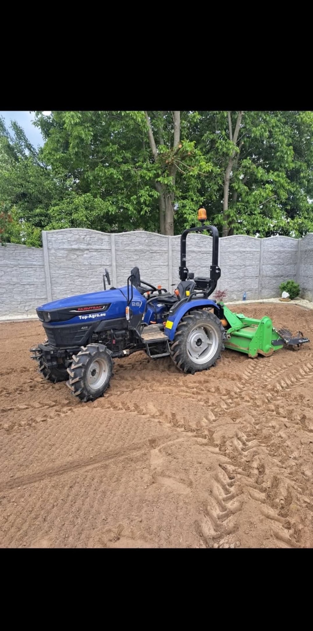 Niebieski traktor Farmtrac z zielonym glebogryzem na przygotowanym, wyrównanym terenie pod przyszły ogród, widoczny betonowy płot i drzewa w tle.