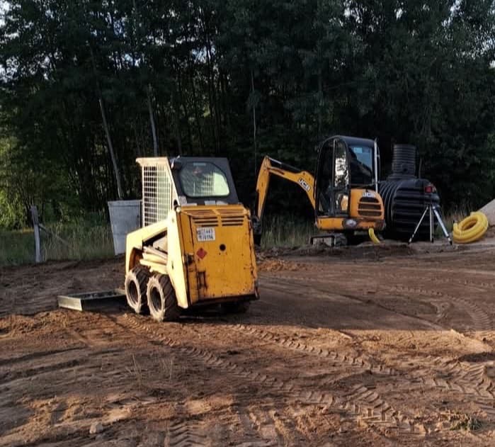 Żółty miniładowarka i koparka JCB na placu budowy, w tle widoczna czarna przydomowa oczyszczalnia ścieków z rurą. Ślady opon na ziemi.