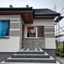 Dwukondygnacyjny dom z szarą elewacją w paski, kamiennym wykończeniem wejścia i czarnym dachem, widoczne rusztowanie z boku.