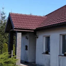 Dom jednorodzinny z nowym, brązowym dachem z blachodachówki, widoczne elementy elewacji, lampy zewnętrzne oraz roślinność w ogrodzie.