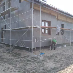 Dom w trakcie ocieplania szarym styropianem, widoczne rusztowania i pracownik w zielonej koszulce przy ścianie.