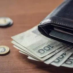 Otwarty skórzany portfel z wystającymi banknotami 100 zł i kartami płatniczymi, obok leżą monety na drewnianym biurku.