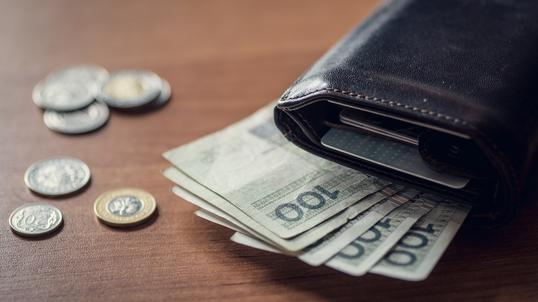 Otwarty skórzany portfel z wystającymi banknotami 100 zł i kartami płatniczymi, obok leżą monety na drewnianym biurku.