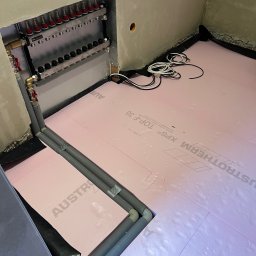 Termico - Rozdzielacz i rury instalacji ogrzewania podłogowego na izolacji z różowego styropianu. Widoczne przewody elektryczne. Ściany w surowym stanie.