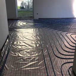 Wnętrze w trakcie montażu ogrzewania podłogowego: rury grzewcze w kolorze niebieskim ułożone na folii z czerwoną siatką, widok z niskiej perspektywy, w tle okno z widokiem na ogród.