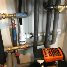 Szczegółowe ujęcie instalacji grzewczej z rurami izolowanymi, zaworami, manometrem i etykietami opisującymi poszczególne elementy, obok leży drukarka etykiet Brother P-touch.