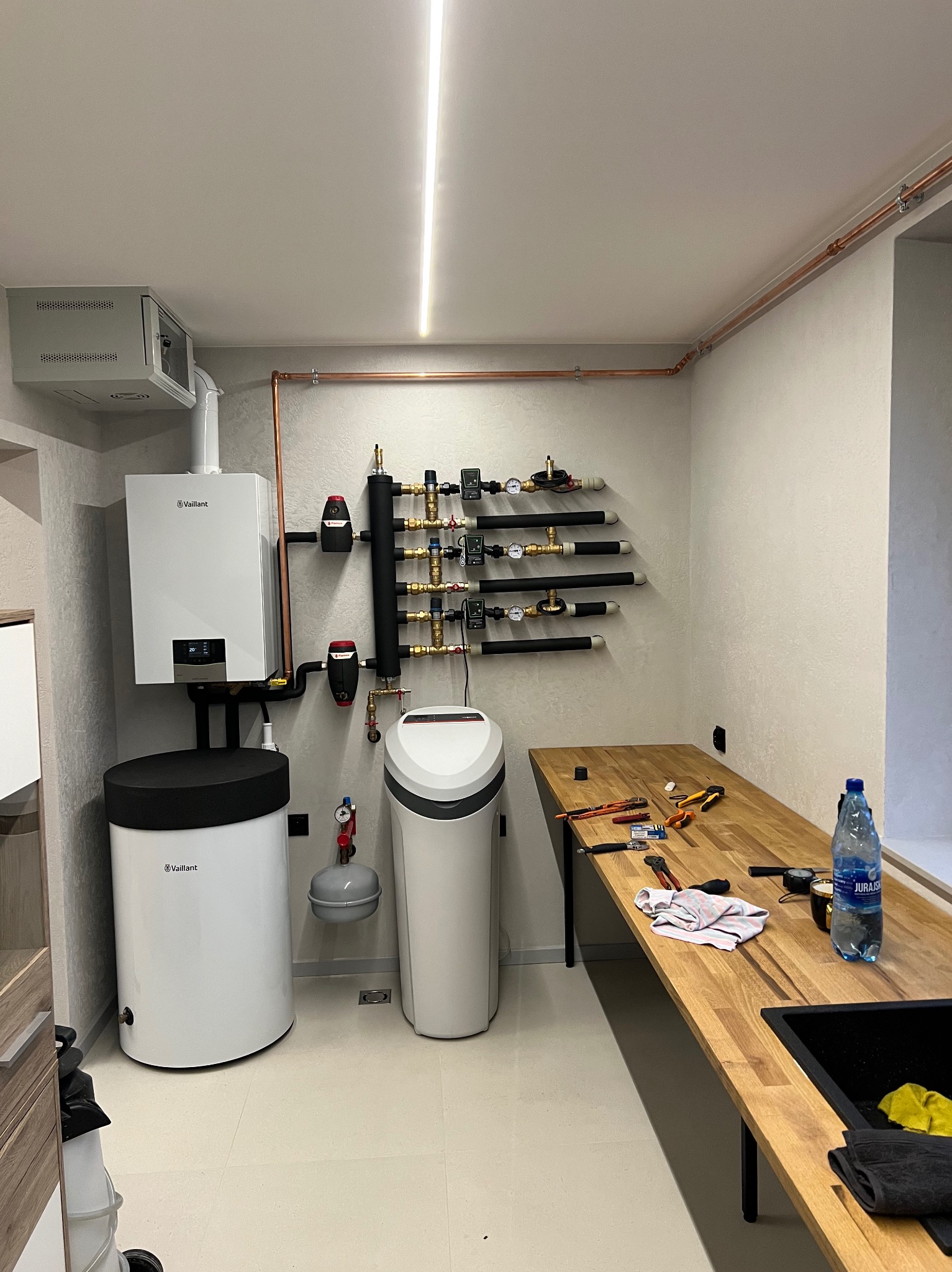 Nowoczesna kotłownia z białym piecem Vaillant, zmiękczaczem wody i miedzianą instalacją. Na drewnianym blacie narzędzia hydrauliczne i butelka wody.