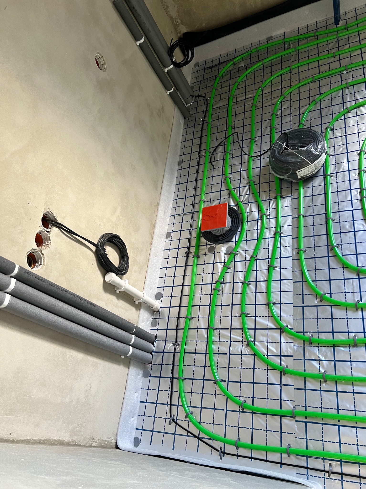 Zielone rury ogrzewania podłogowego ułożone na izolacji z folią w kratkę, widoczne kable i rury przy ścianie. Etap instalacji przed wylewką.