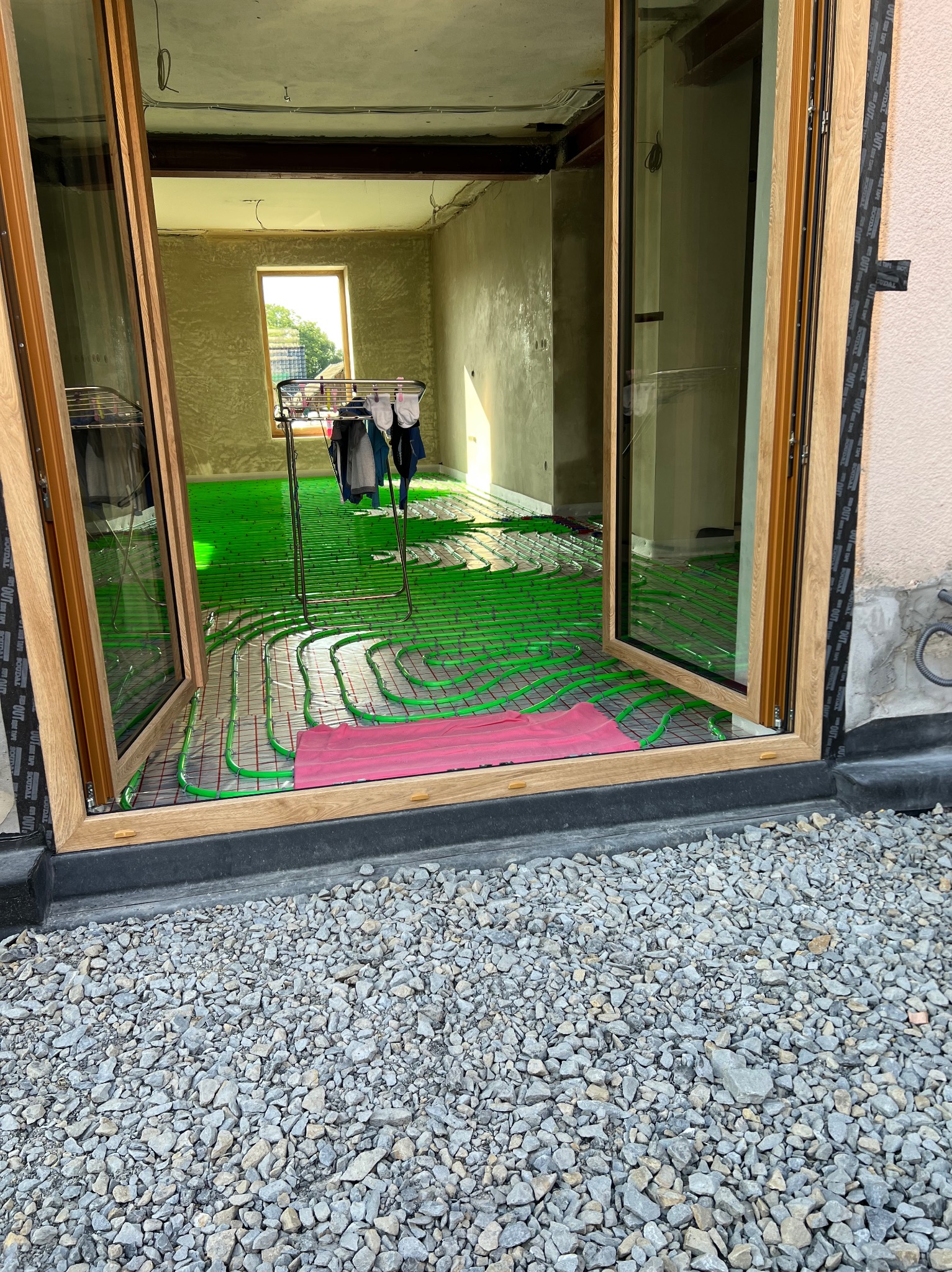 Zielone rury ogrzewania podłogowego ułożone w pokoju, widoczne przez otwarte drzwi balkonowe. Na rurach leży różowy ręcznik i suszarka z ubraniami.