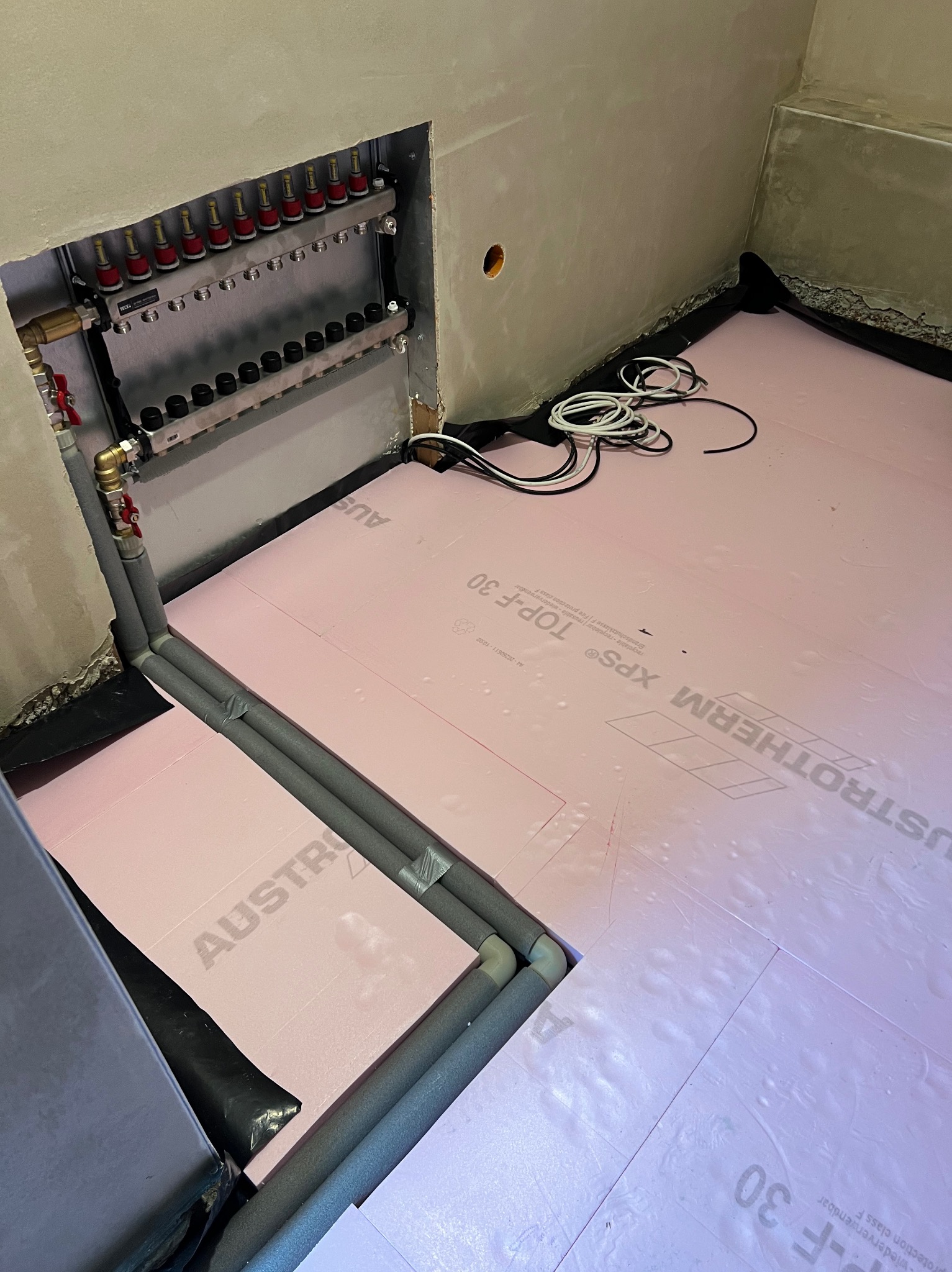Rozdzielacz i rury instalacji ogrzewania podłogowego na izolacji z różowego styropianu. Widoczne przewody elektryczne. Ściany w surowym stanie.