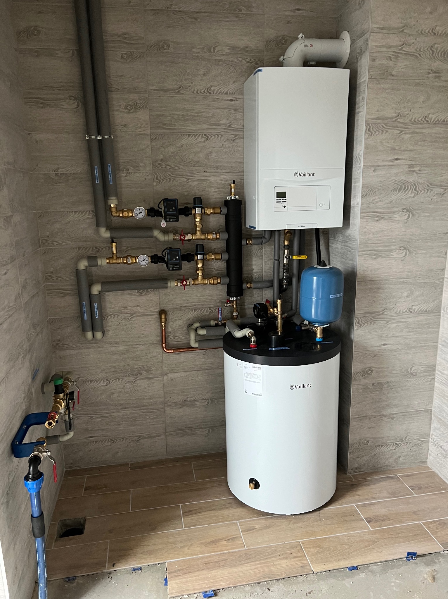 Biały piec gazowy Vaillant z szarymi rurami i niebieskim zbiornikiem wyrównawczym na tle płytek imitujących drewno. Instalacja w kotłowni.