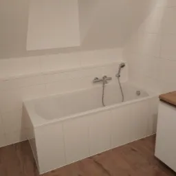 Biała wanna obudowana płytkami w łazience na poddaszu, bateria i słuchawka prysznicowa zamontowane na ścianie, drewnopodobna podłoga, szafka z drewnianym blatem po prawej stronie.