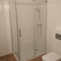 Kabina prysznicowa z przesuwnymi drzwiami ze srebrnymi okuciami, zamontowana w łazience z beżowymi płytkami i drewnopodobną podłogą, widoczna toaleta podwieszana.