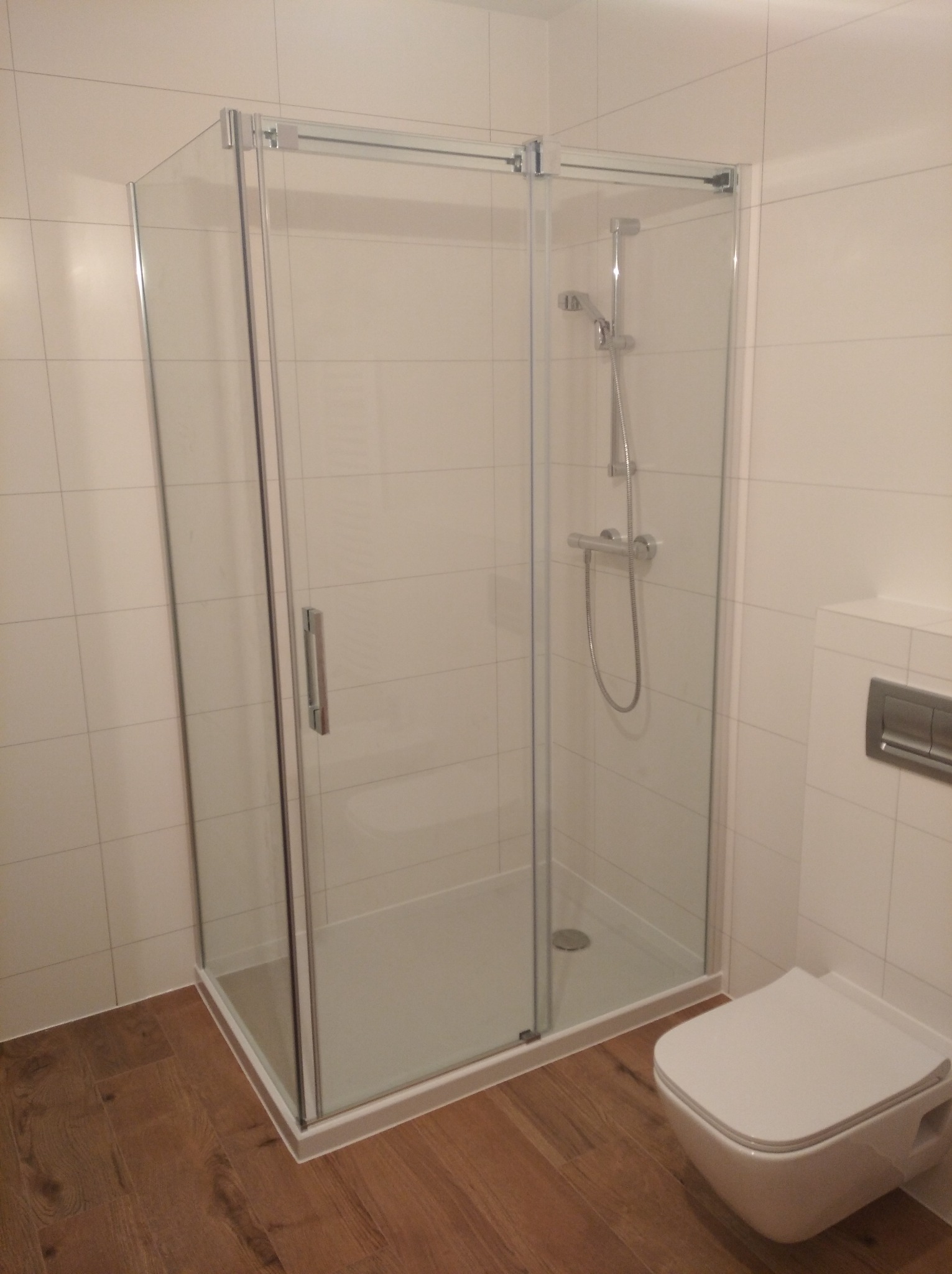 Kabina prysznicowa z przesuwnymi drzwiami ze srebrnymi okuciami, zamontowana w łazience z beżowymi płytkami i drewnopodobną podłogą, widoczna toaleta podwieszana.