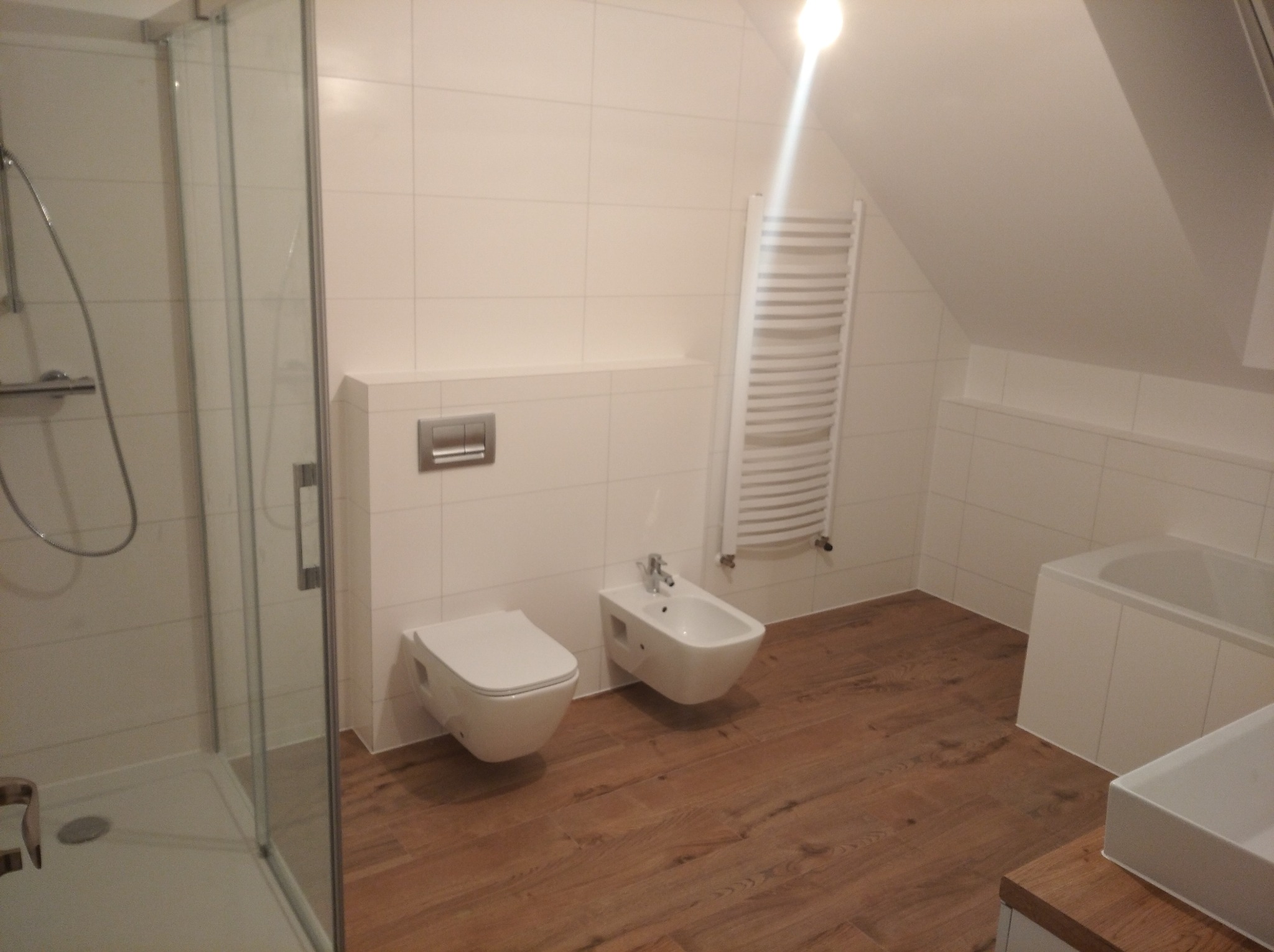 Nowoczesna łazienka na poddaszu z drewnopodobną podłogą, białymi płytkami, kabiną prysznicową, wiszącą toaletą i bidetem oraz grzejnikiem drabinkowym.