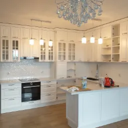 Jasna kuchnia z białymi szafkami, wyspą i kryształowym żyrandolem, widok na płytę indukcyjną i srebrną lodówkę.