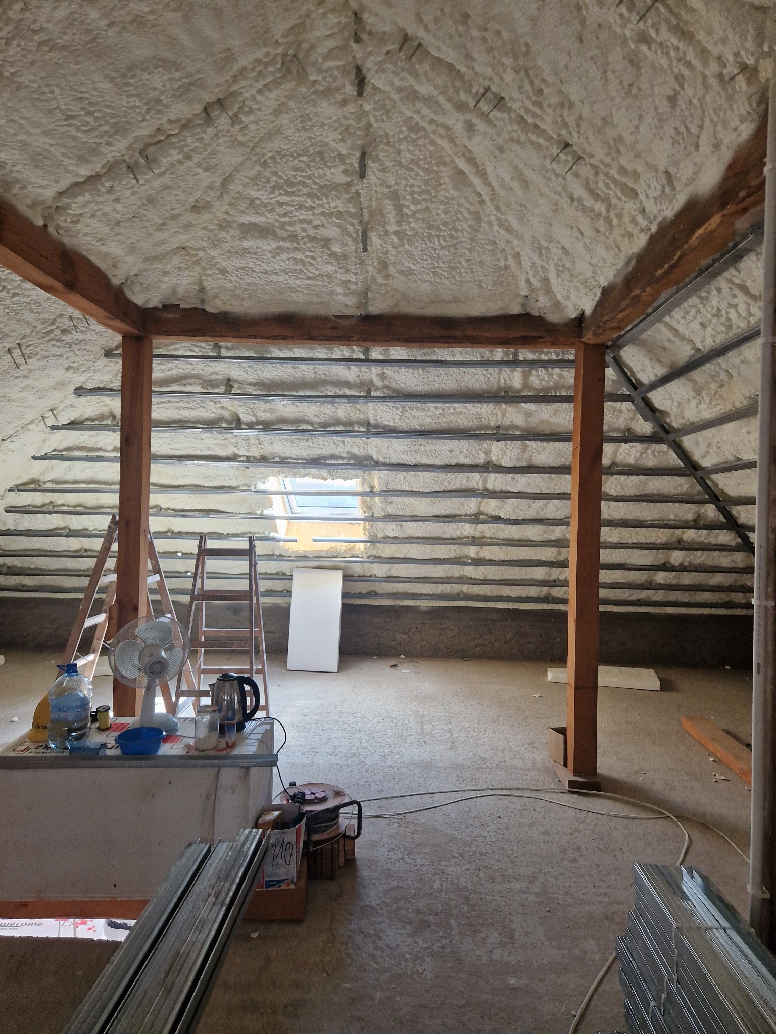 Poddasze w trakcie prac izolacyjnych, pokryte pianą natryskową, widoczne drewniane belki konstrukcyjne, drabiny, wentylator oraz materiały budowlane na podłodze.