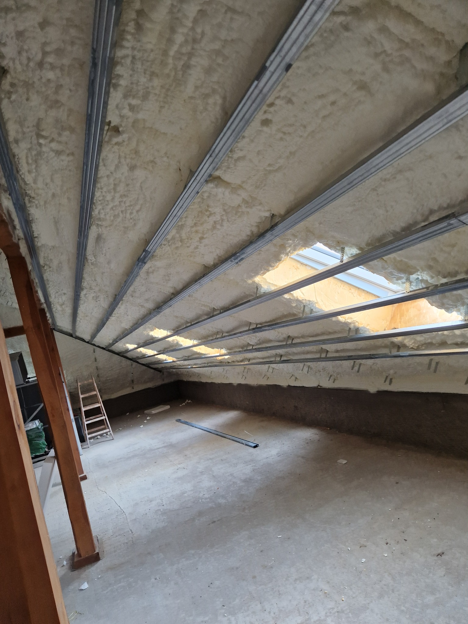 Poddasze z widoczną izolacją natryskową z pianki poliuretanowej między metalowymi profilami konstrukcyjnymi, częściowo wykończone, z oknami dachowymi.