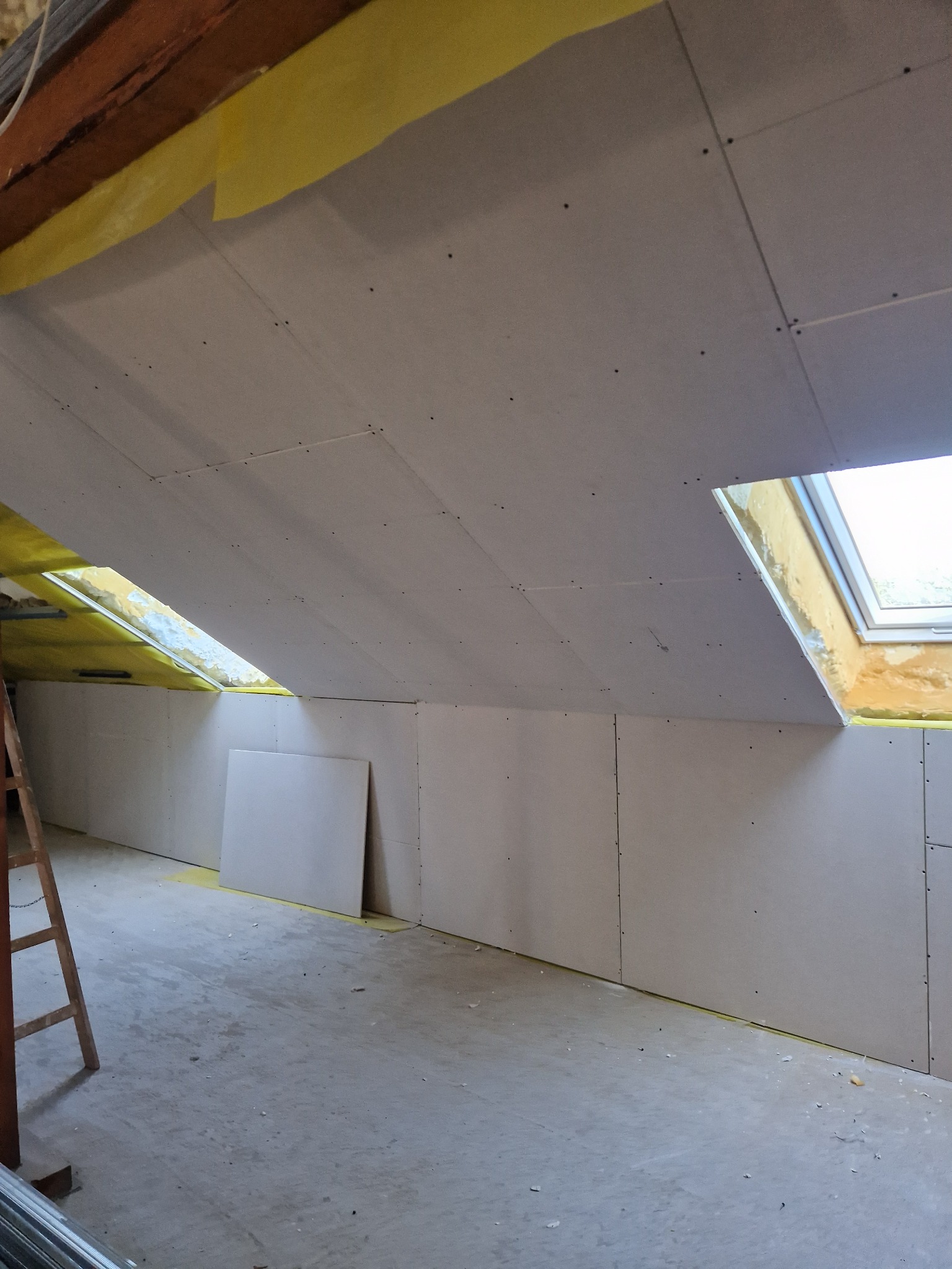 Poddasze z oknami dachowymi w trakcie zabudowy: widoczne płyty gipsowo-kartonowe, szpachlowane łączenia, częściowo ocieplona przestrzeń poddasza, drabina oparta o ścianę.
