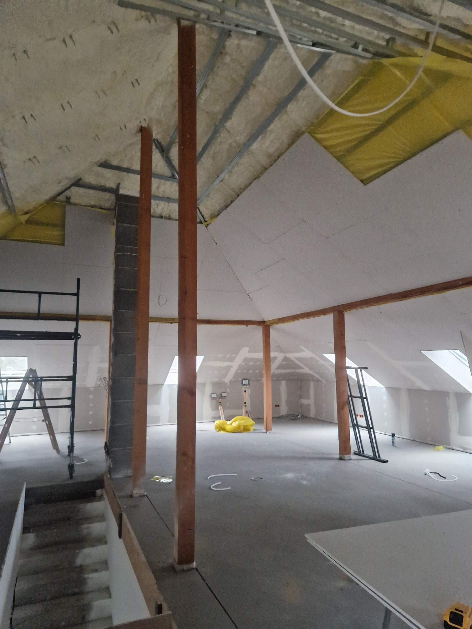 Poddasze w trakcie adaptacji: widoczne drewniane słupy, rusztowanie, częściowo ułożona izolacja, płyty gipsowe na suficie, przygotowane do dalszych prac wykończeniowych.