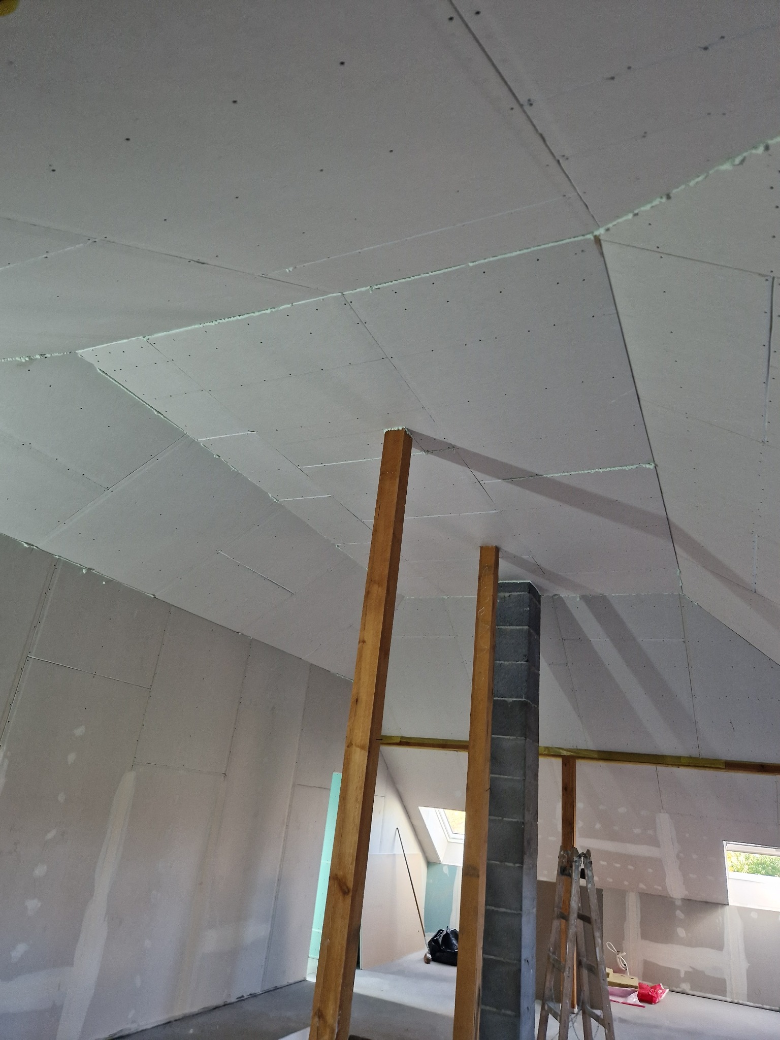 Skośne poddasze w trakcie prac wykończeniowych, widoczne płyty na suficie i ścianach, drewniane podpory i komin z cegły, drabina oparta o ścianę.