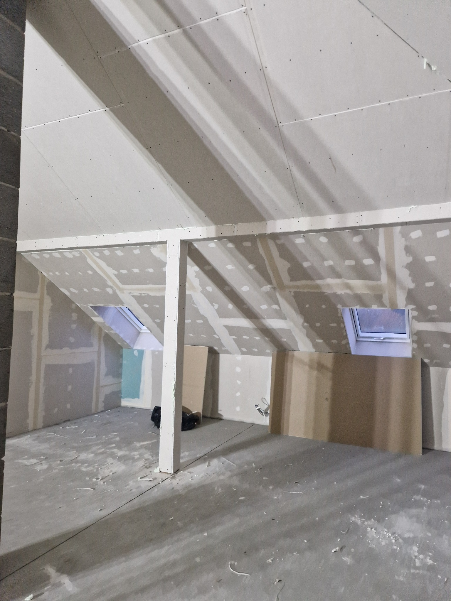 Poddasze w trakcie prac wykończeniowych: zamontowane płyty na skosach i suficie, widoczne okna dachowe, ślady po szpachlowaniu na ścianach, surowy beton na podłodze, słup podtrzymujący konstrukcję.