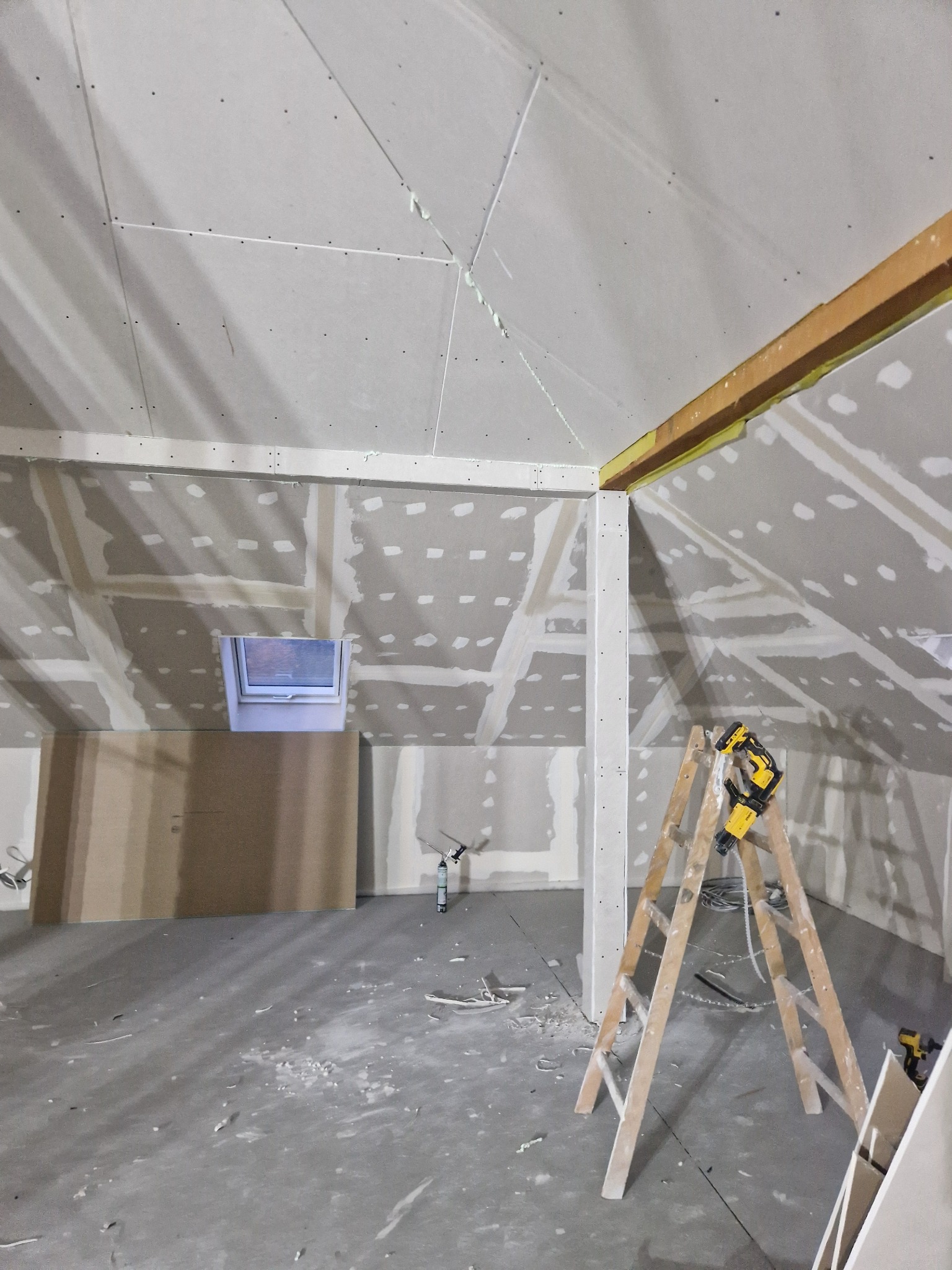 Poddasze w trakcie prac wykończeniowych, widoczne płyty na skosach, szpachlowane łączenia, okno dachowe, drabina z narzędziami i resztki materiałów na podłodze.