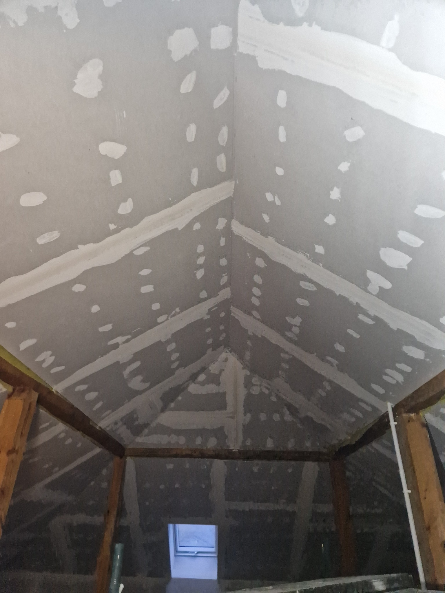 Skośny sufit poddasza z płyt kartonowo-gipsowych w trakcie szpachlowania łączeń, widoczne drewniane elementy konstrukcyjne i okno dachowe w dolnej części kadru.