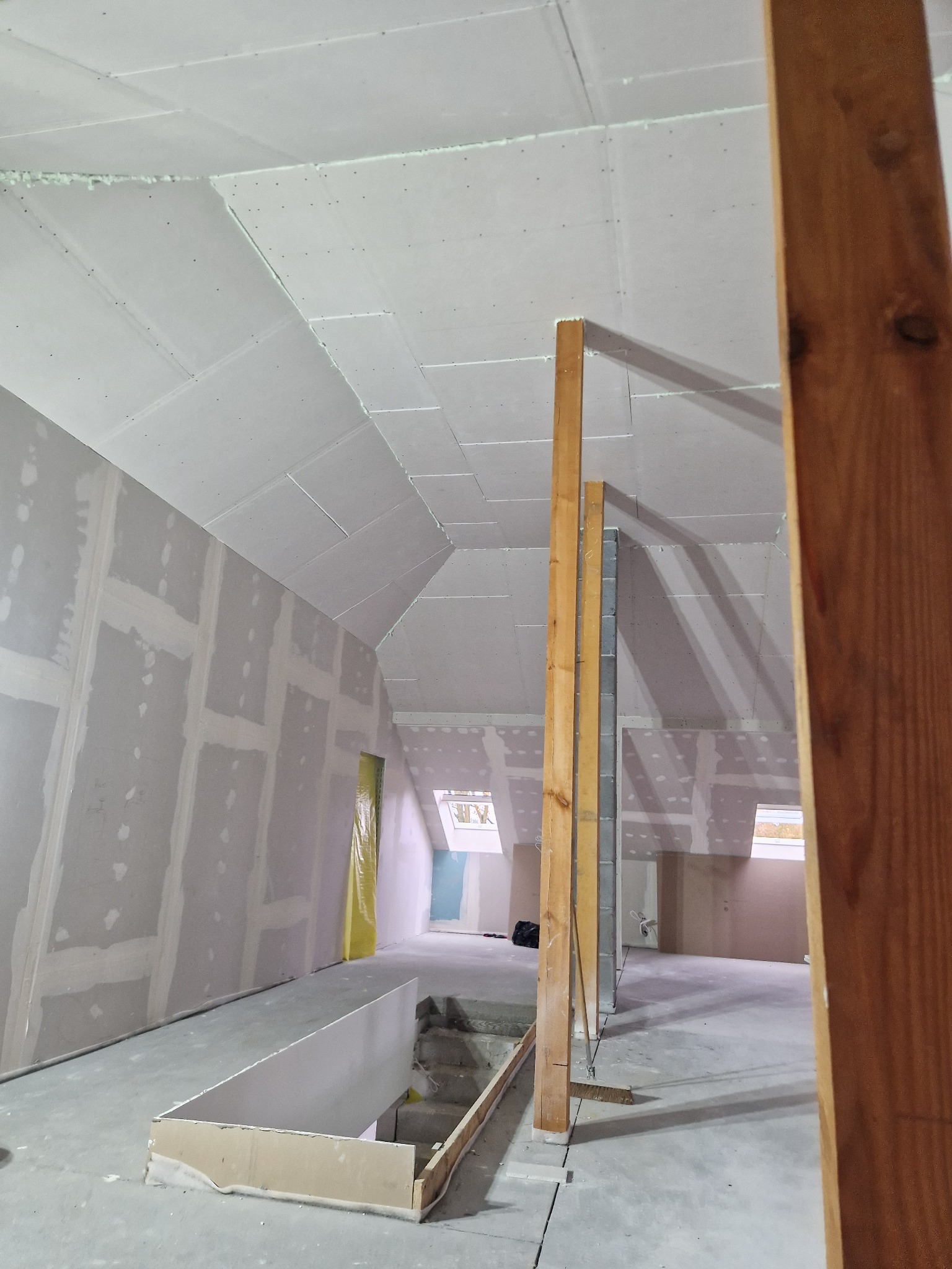 Poddasze w trakcie adaptacji: widoczne drewniane stemple podtrzymujące konstrukcję, ściany i skosy obłożone płytami, łączenia płyt częściowo zaszpachlowane, surowe wykończenie podłogi.