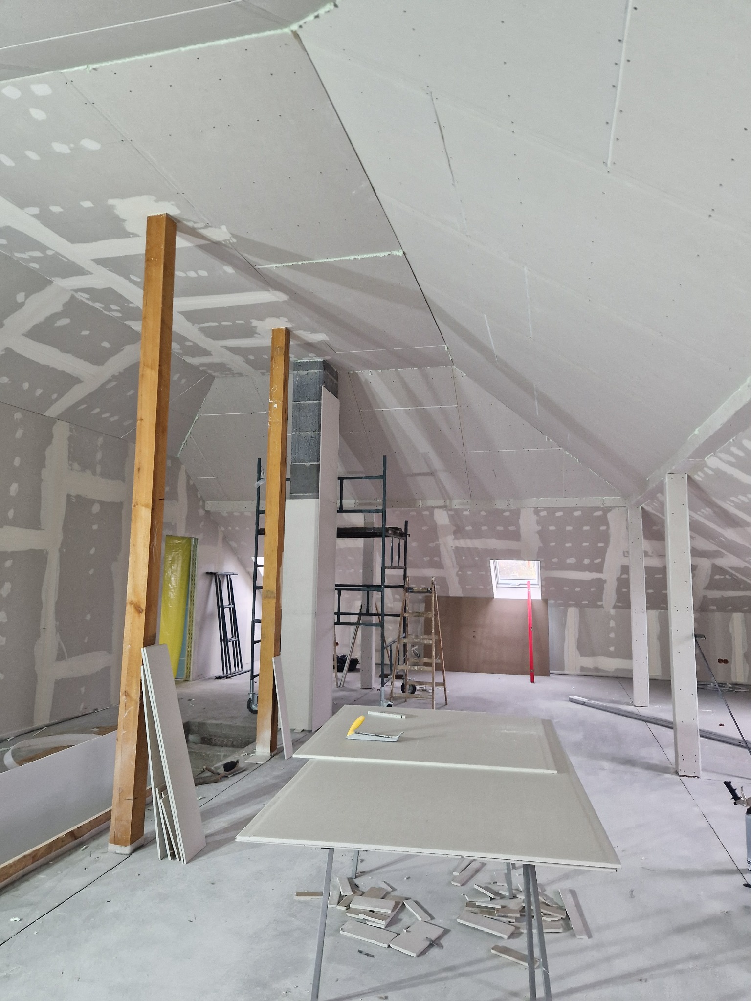Poddasze w trakcie prac wykończeniowych: montaż płyt na skośnym suficie, widoczne drewniane podpory, rusztowania i narzędzia, przygotowane płyty na stole roboczym, surowy stan ścian.