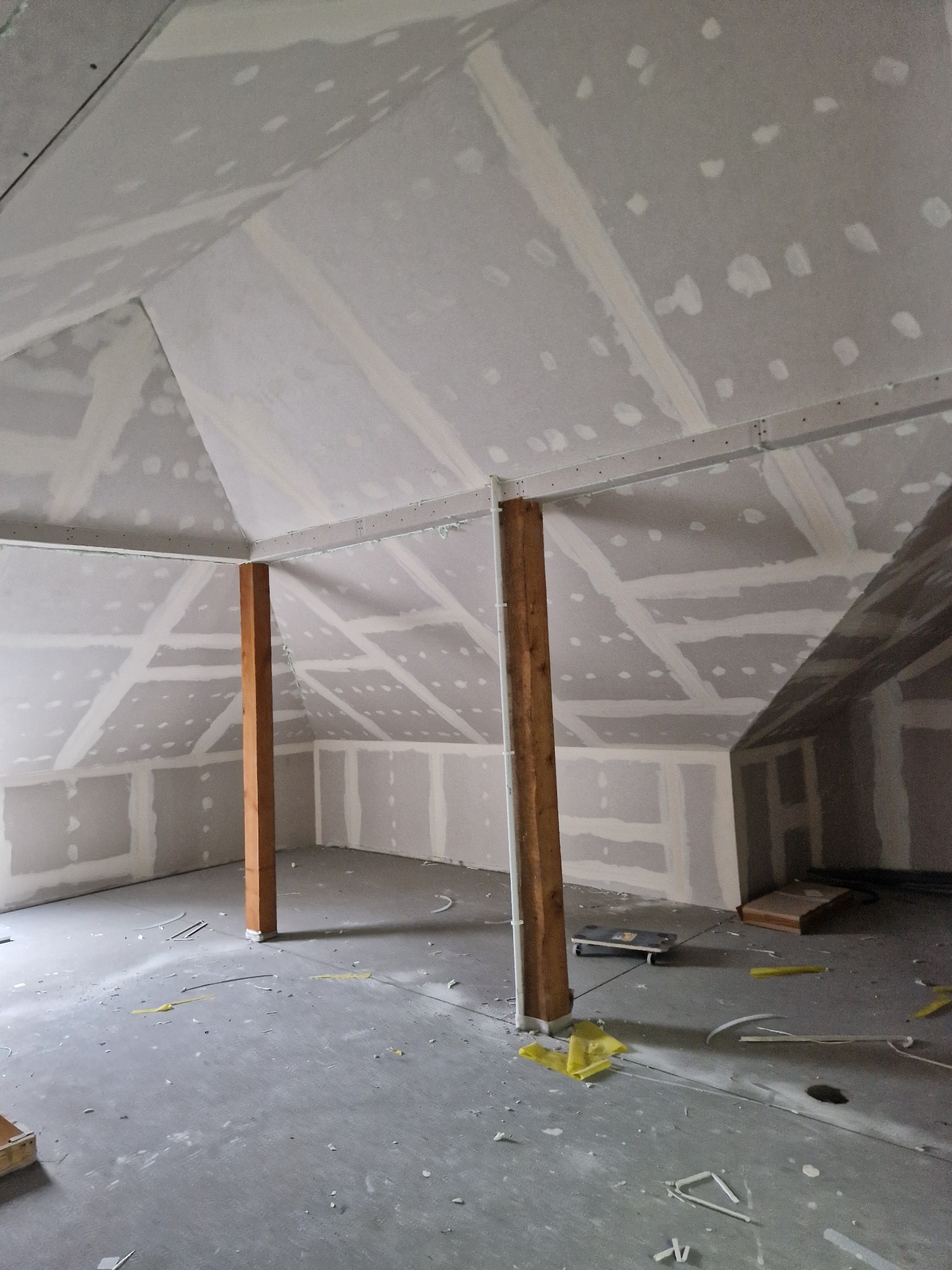 Poddasze w trakcie wykańczania: zamontowane płyty gipsowo-kartonowe na skośnych ścianach i suficie, widoczne drewniane słupy podtrzymujące konstrukcję, ślady po szpachlowaniu na łączeniach płyt...