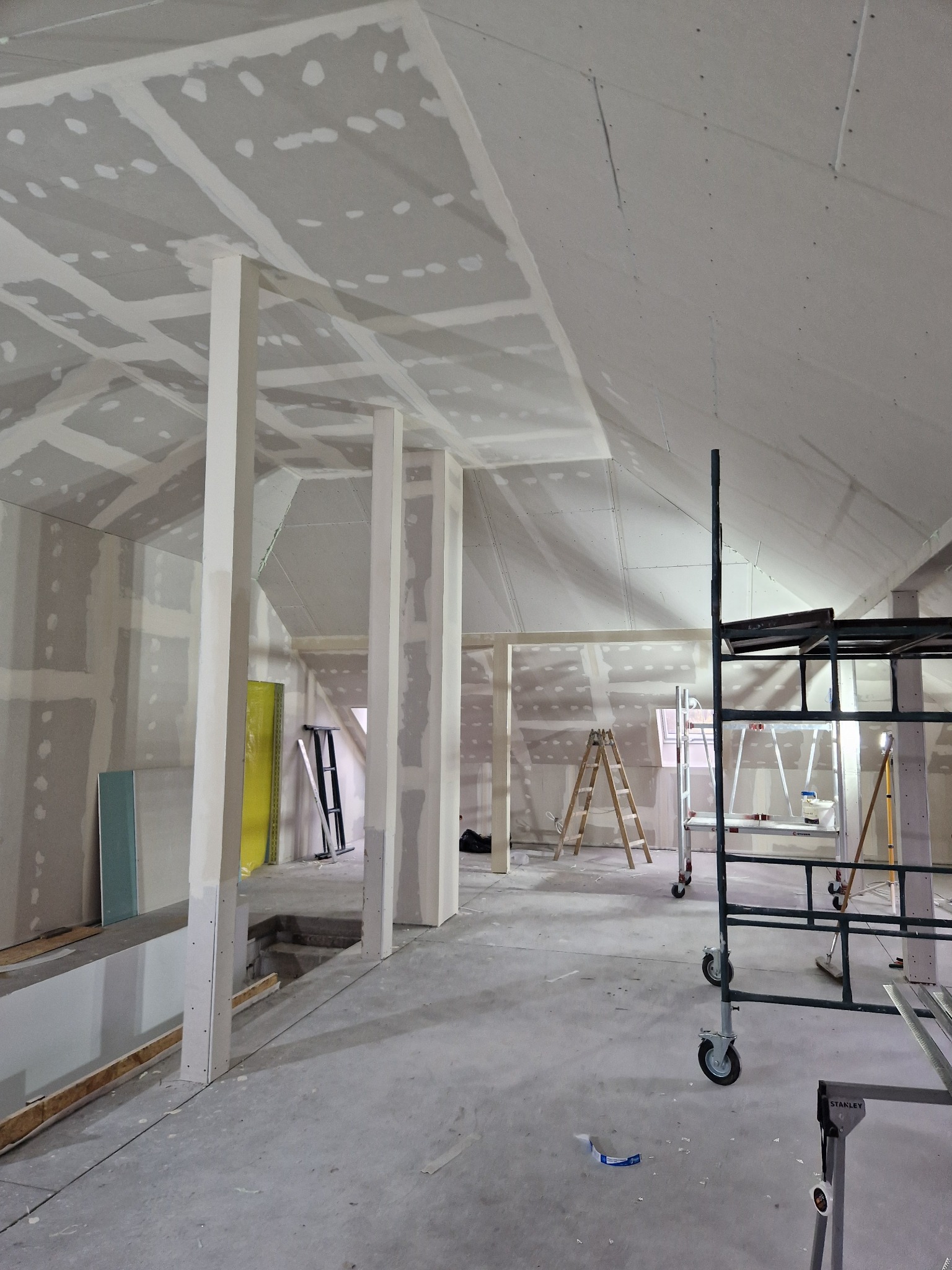 Poddasze w trakcie wykańczania płytami kartonowo-gipsowymi, widoczne łączenia płyt wypełnione szpachlą, drewniane i metalowe rusztowania w tle, przygotowane do dalszych prac wykończeniowych.