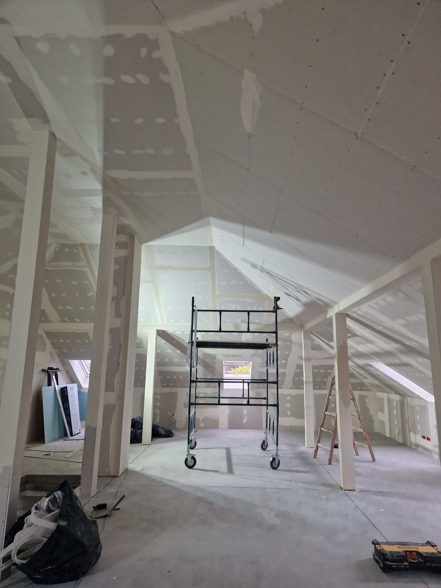 Poddasze w trakcie wykańczania: widoczne stelaże, płyty pokrywające skosy, łączenia płyt przygotowane do dalszej obróbki, rusztowanie na środku, drabina oparta o ścianę.