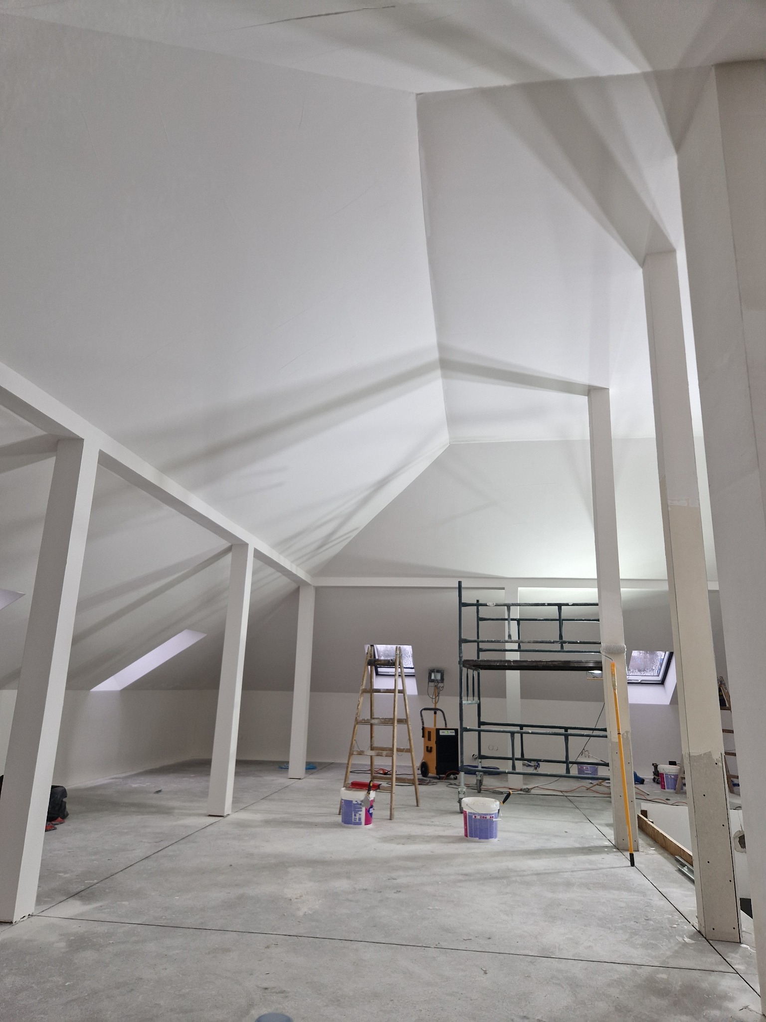 Poddasze w trakcie malowania na biało, widoczne rusztowanie, drabina, wiaderka z farbą i wałek oparty o filar, w tle okna dachowe.