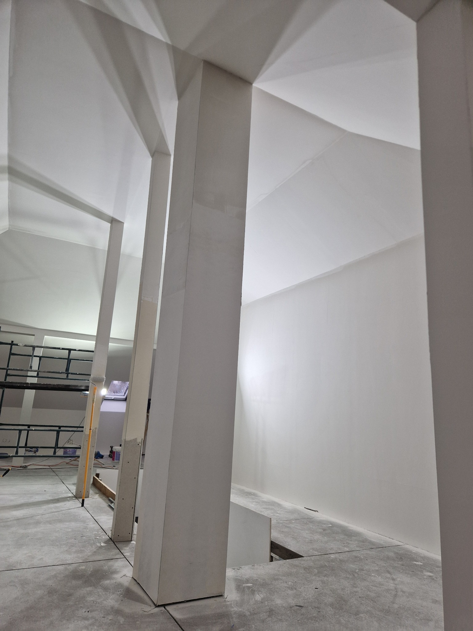 Wnętrze w trakcie wykańczania, widoczne pionowe słupy konstrukcyjne obłożone płytami, surowa podłoga, w tle rusztowanie i ściany przygotowane do dalszych prac wykończeniowych.